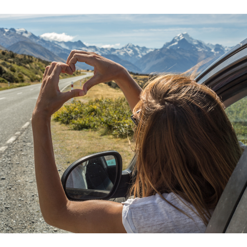 Young woman leaning out car window as she's pulled over, making a heart with her hands. Mountains in the distance.
