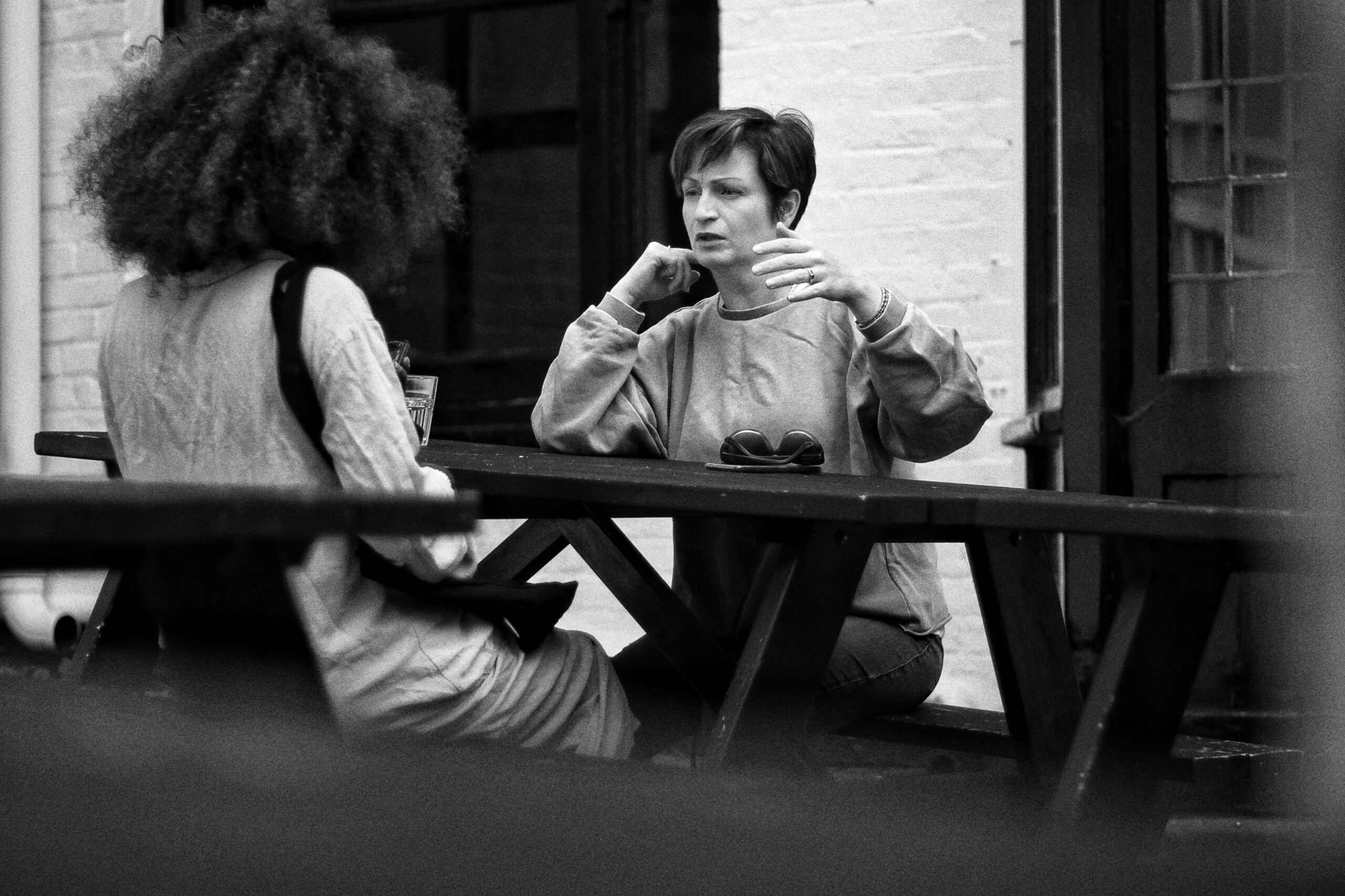 Two people at a picnic table outside a building have a conversation