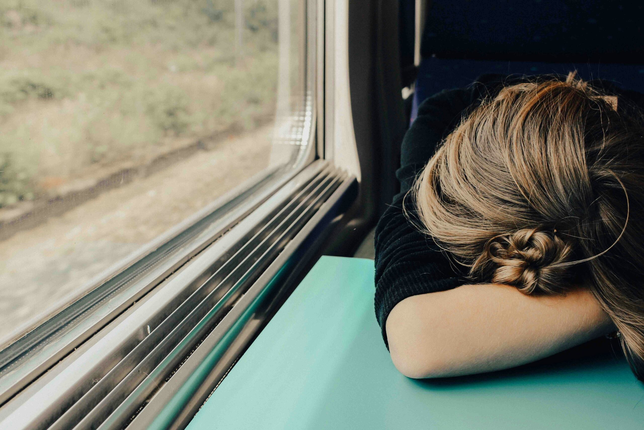 A young woman is sleeping on a train, her head resting against the window as the sun sets outside. Her relaxed posture illustrates a deep sense of fatigue and the exhaustion that often accompanies daily life. The soft glow of the sunset adds warmth to the scene, highlighting the challenges of finding rest amidst the hustle and bustle of commuting, especially during the time change of daylight savings.