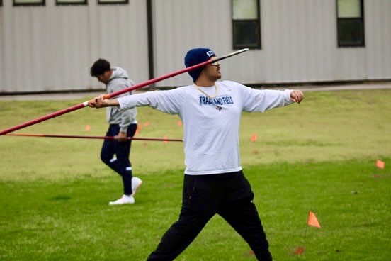 A collegiate athlete practicing javelin throw on a grassy field, wearing a white long-sleeve shirt and a blue cap, with another person practicing in the background.