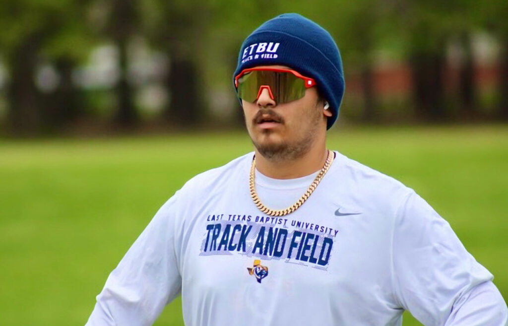 A collegiate track and field athlete wearing a beanie and sunglasses, standing on a field in a white athletic shirt with 'East Texas Baptist University Track and Field' printed on it.
