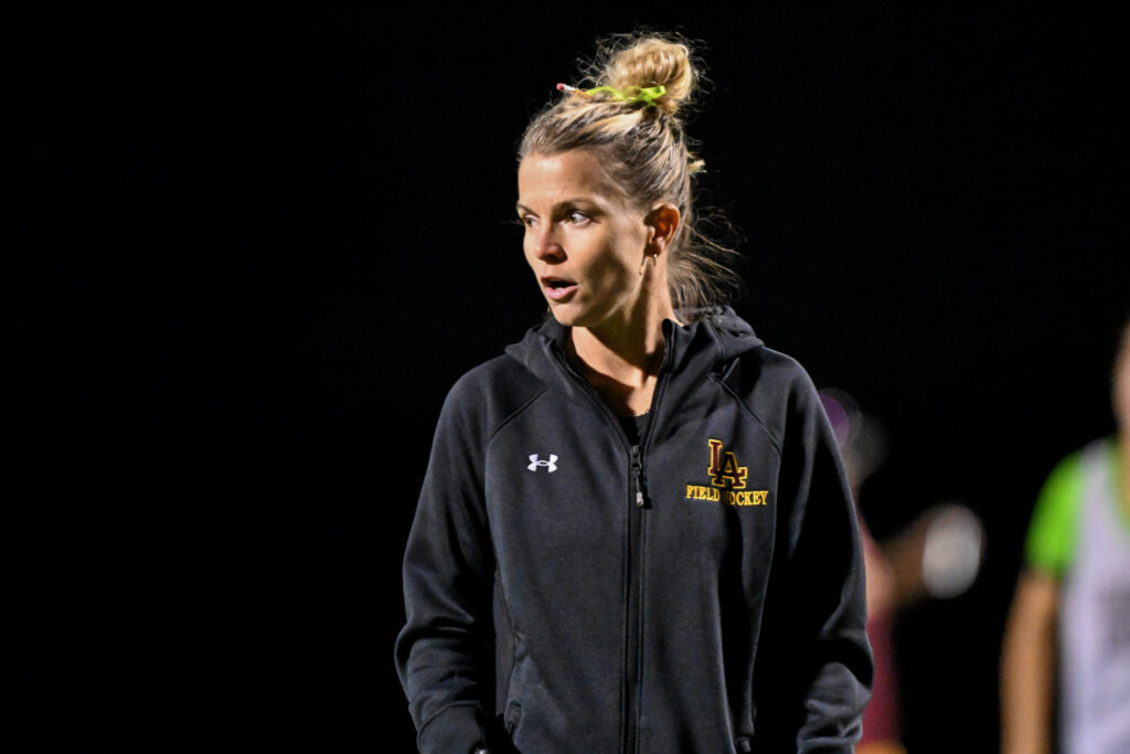A female field hockey coach in a black jacket, looking focused while observing a training session at night.