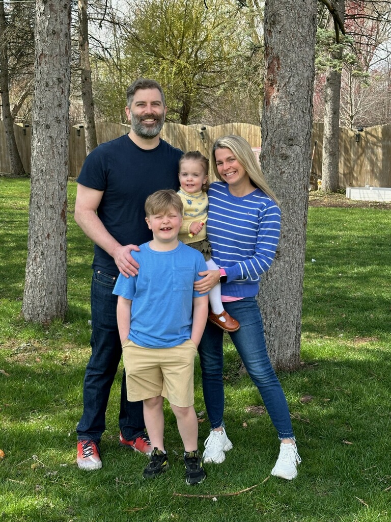 A family of four poses together outdoors, smiling. The father stands on the left wearing a black t-shirt and dark pants, while the mother, on the right, wears a striped blue and white sweater. The son, in a blue shirt and beige shorts, stands in front of them, and the daughter, dressed in a yellow outfit, is held by the mother.