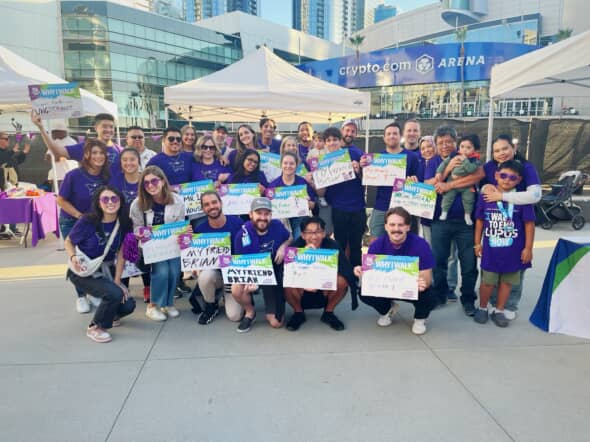 Group photo of individuals wearing purple shirts, gathered outdoors at an event promoting lupus awareness, holding signs and smiling together.